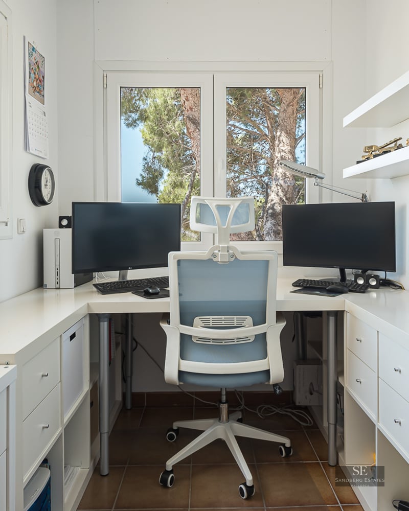 Modern white desk with dual monitors, ergonomic chair, and windows overlooking green trees.