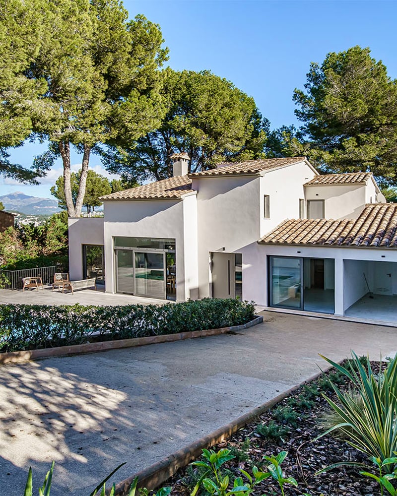 A two-story modern white villa with terracotta roof tiles, surrounded by tall pine trees and a swimming pool area.