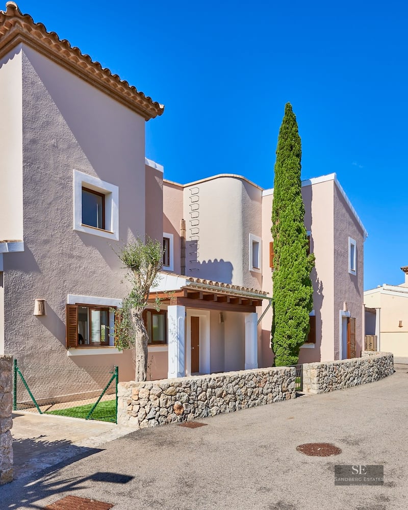 Mediterranean-style villa in pale terracotta with stone walls, wooden shutters, and a tall cypress tree under a blue sky.