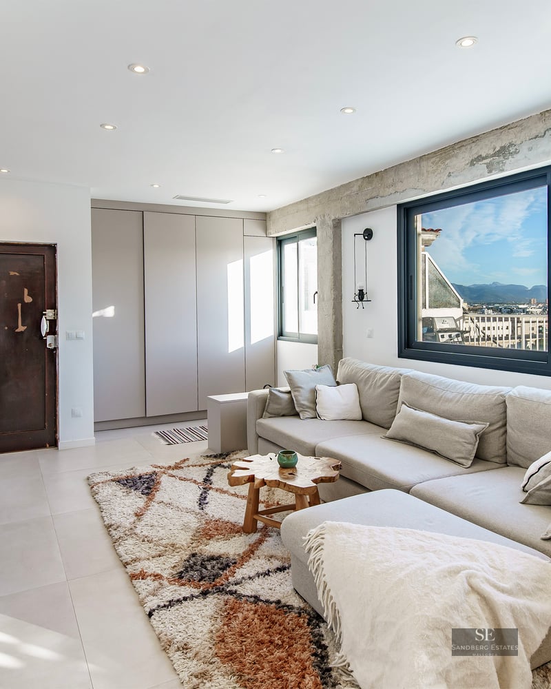 Bright living room with a large grey sofa, exposed concrete beams, and a wide window overlooking a city skyline.