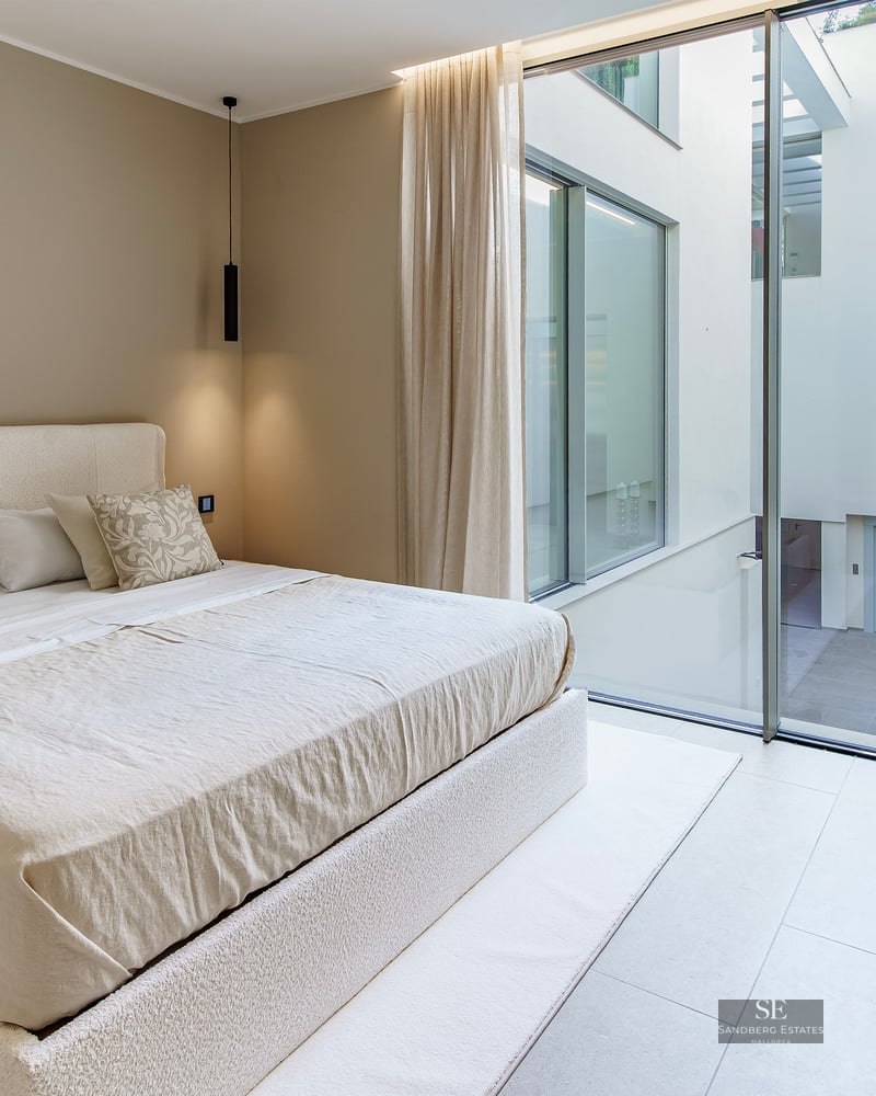 A bright, minimalist master bedroom with a cream boucle bed, matching armchair, and large windows facing an internal courtyard.