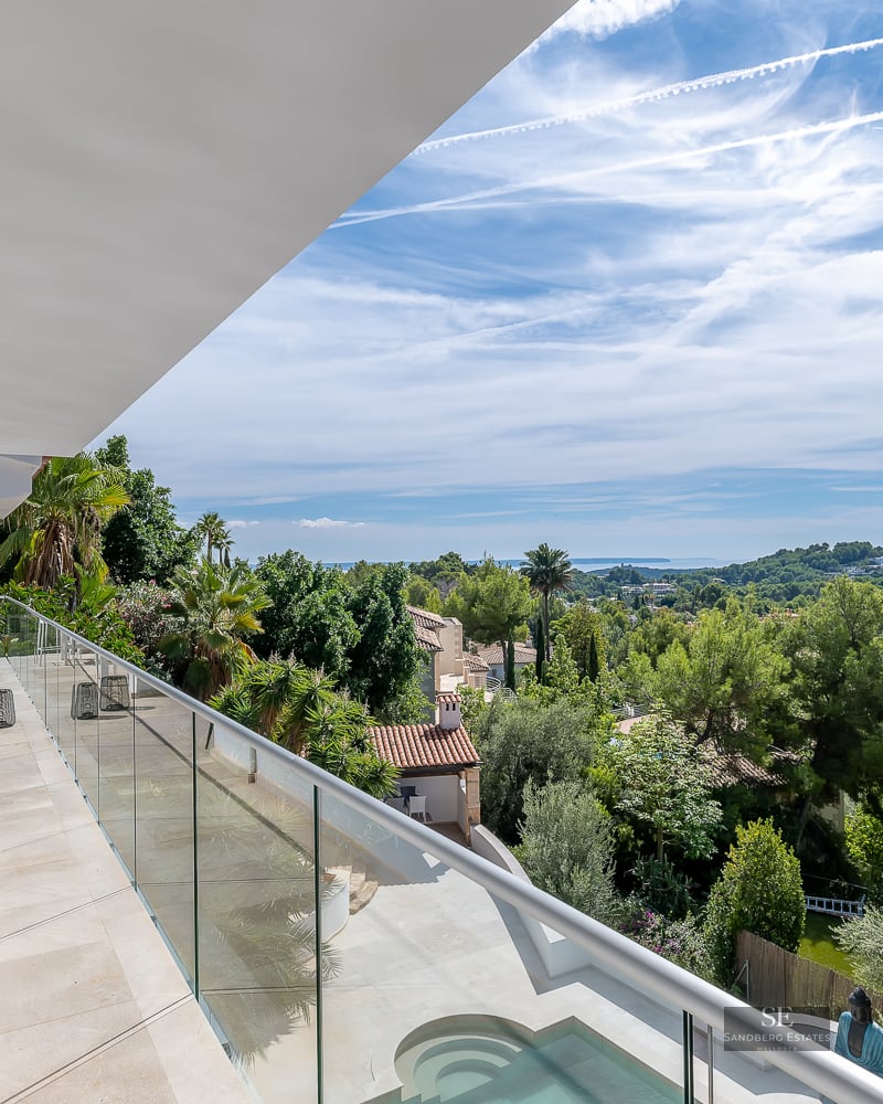 Balcony with glass railing overlooking lush green trees, a swimming pool, and the Mediterranean sea.