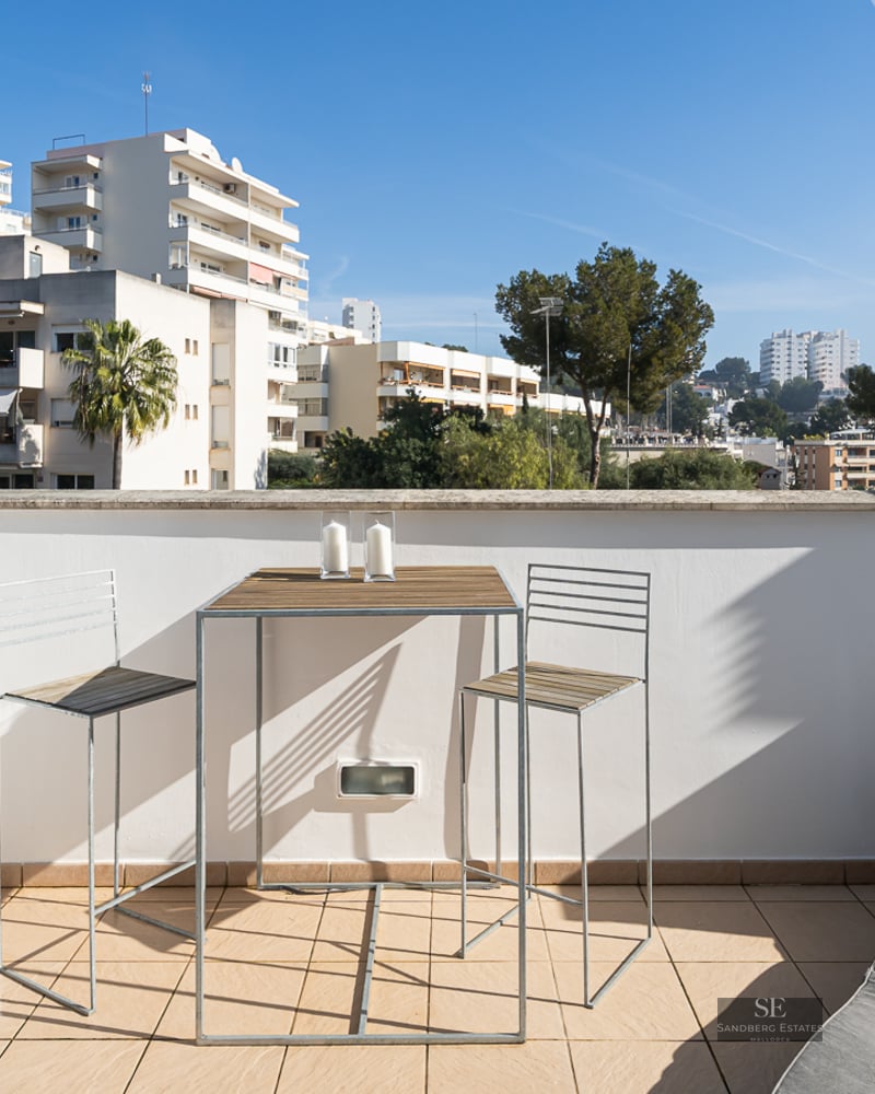 A sunlit terrace with a high-top wooden table, two bar chairs, and a grey sun lounger overlooking city buildings.
