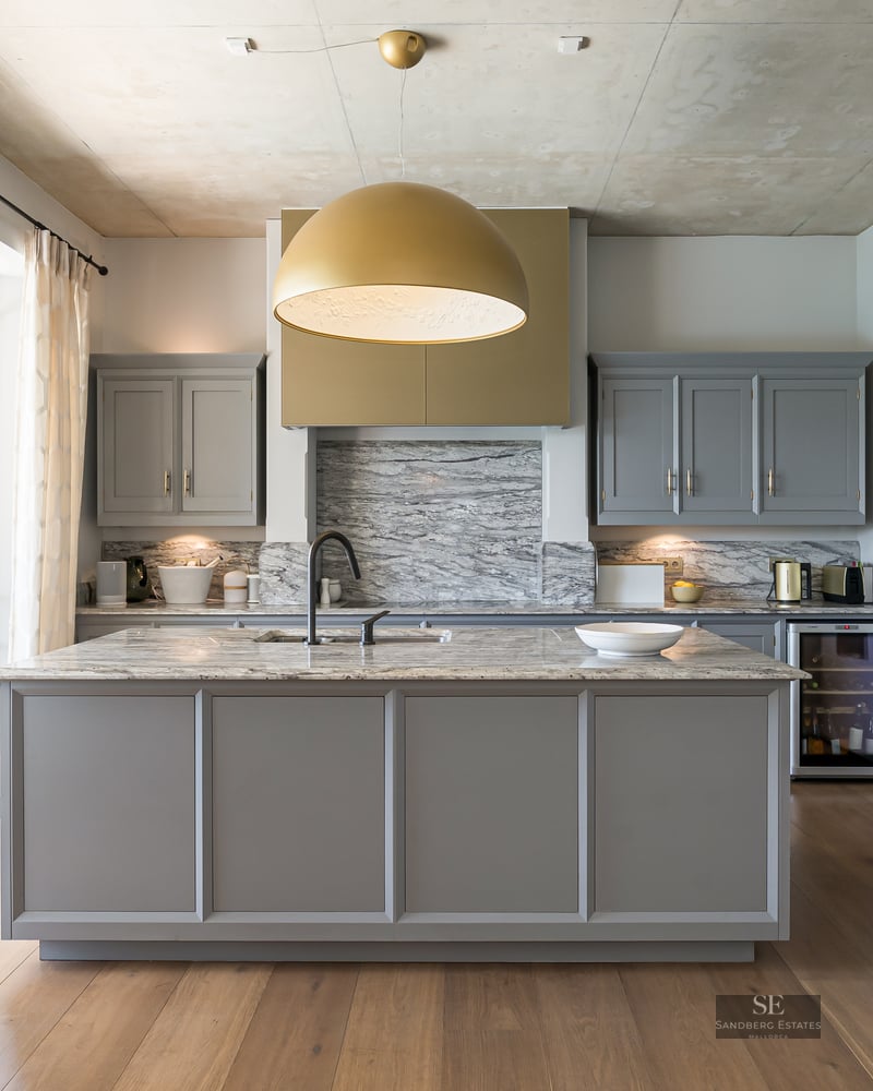 Modern kitchen with grey cabinets, marble island, gold pendant light, and floor-to-ceiling windows to a garden.