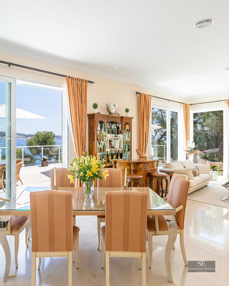 Bright open-plan dining room with a glass table, marble floors, and large windows overlooking the Mediterranean sea.