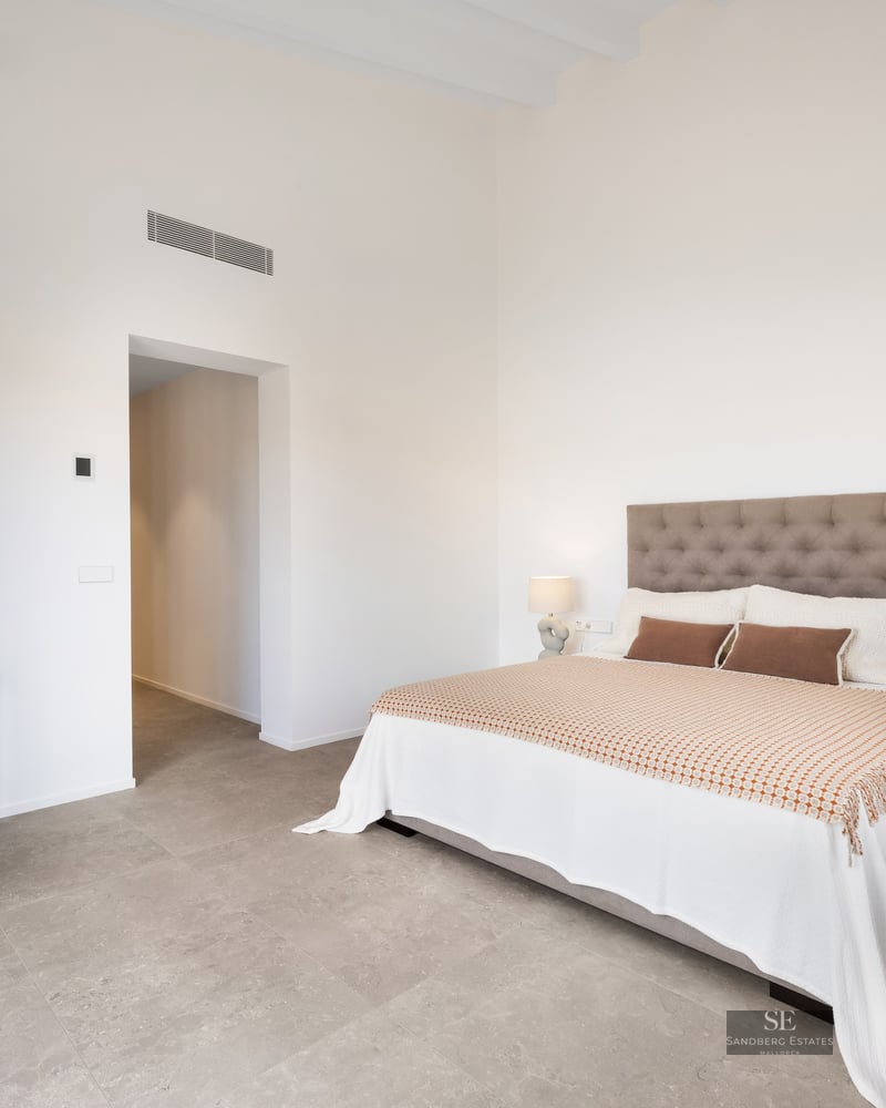 Minimalist bedroom with a tufted headboard, white bedding, designer lamps, and grey stone floors under high ceilings.