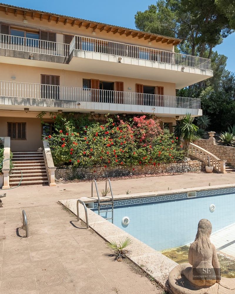 Multi-level Mediterranean villa featuring large terraces and a large empty swimming pool surrounded by lush greenery.