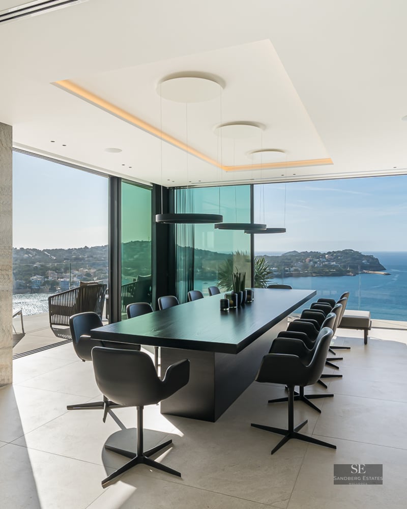 A modern dining room with a long black table, stone fireplace, and floor-to-ceiling windows overlooking the ocean.