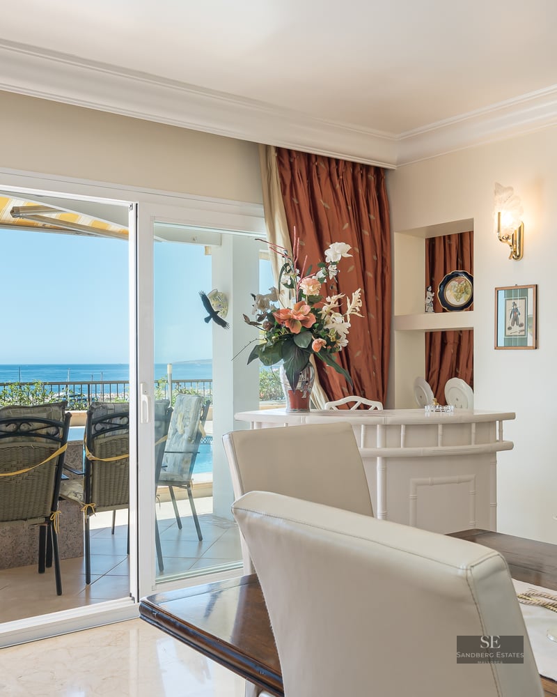 Interior dining area with a wooden table and white chairs looking out to a terrace with a pool and sea views.
