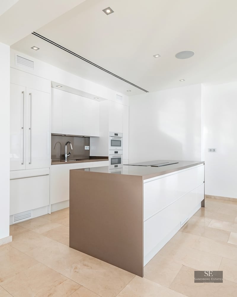 Bright, white modern kitchen featuring a central island, integrated appliances, and sea views through the window.