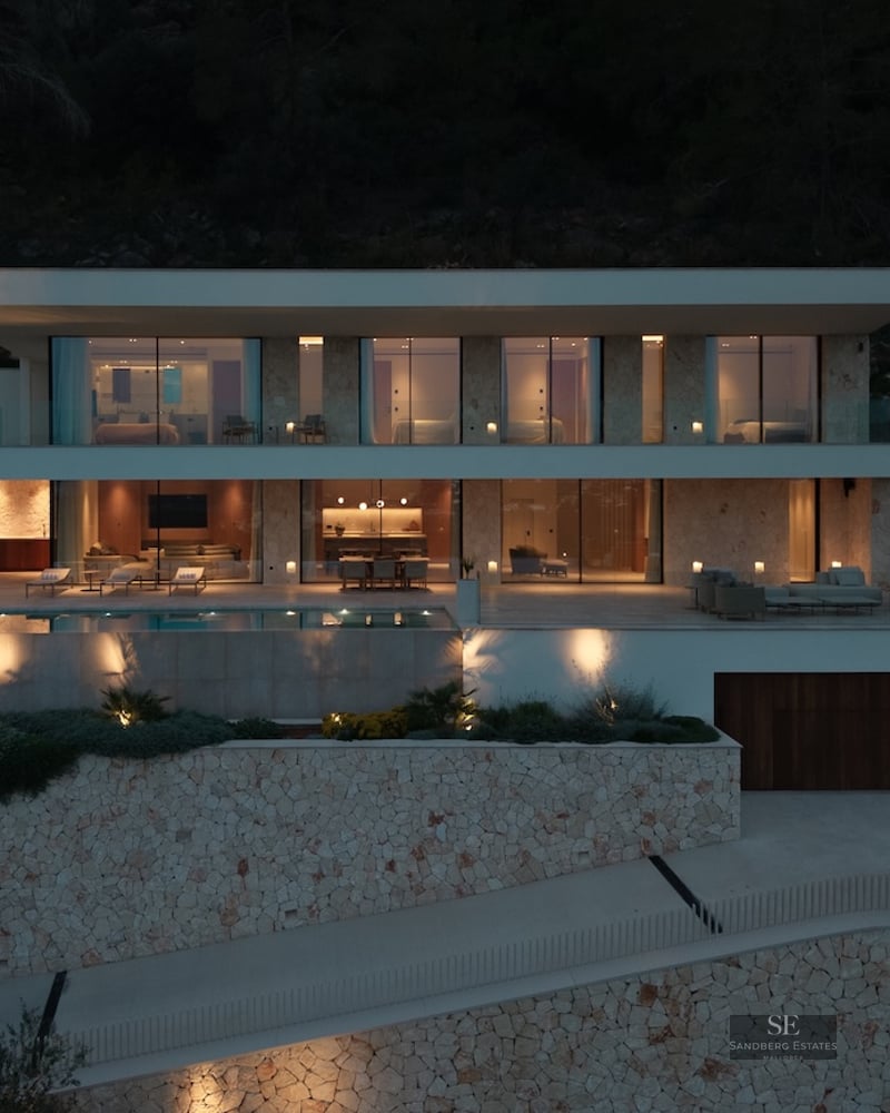 Illuminated nighttime facade of a three-level modern villa featuring an infinity pool and natural stone walls.