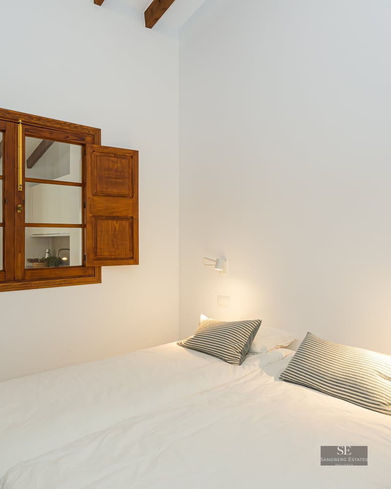 Bright bedroom with white bedding, striped pillows, a traditional wooden window with shutters, and exposed beams.