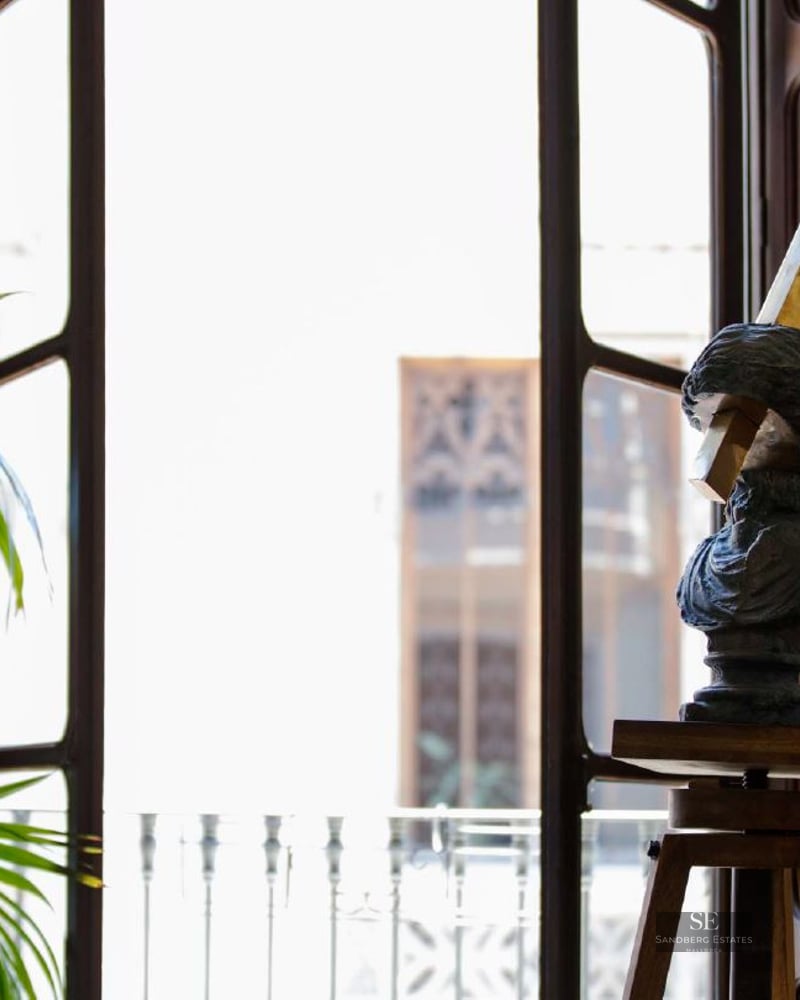 Sculpture of a bust on a wooden tripod stand next to a large bright window with green palm plants.