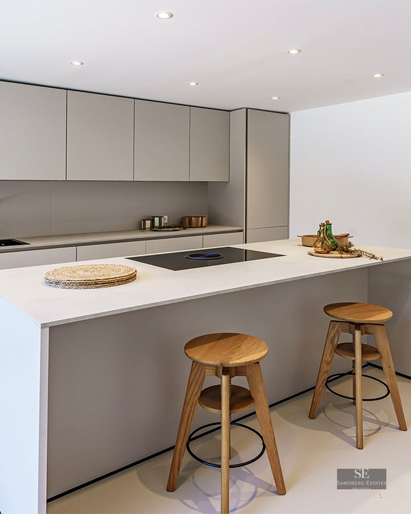Sleek light grey kitchen with a large island, wooden stools, and glass door leading to a terrace.