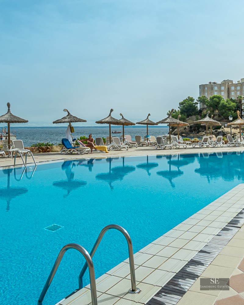 Large blue swimming pool surrounded by sunbeds and straw umbrellas, overlooking the sea and coastal buildings.