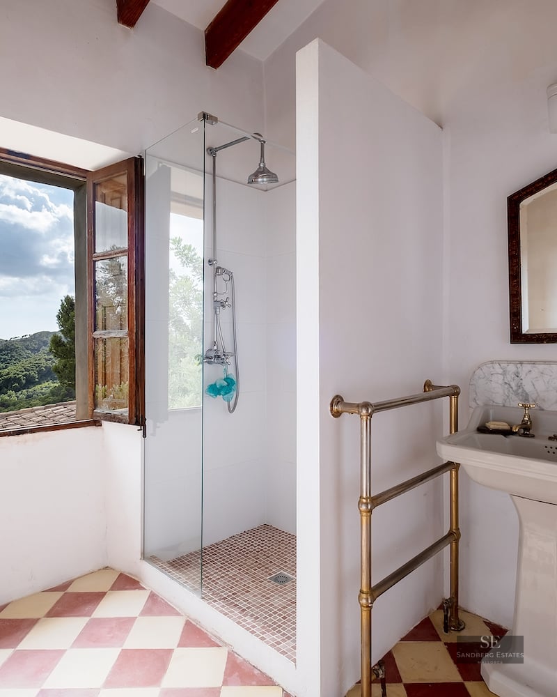 Bathroom with checkered floors, pedestal sink, and glass shower next to a window overlooking green mountains.