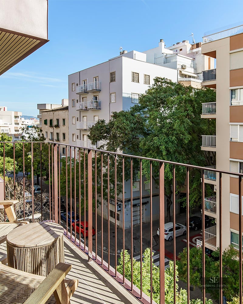 A sunny balcony with wooden chairs and a table overlooking a tree-lined city street and apartment buildings.