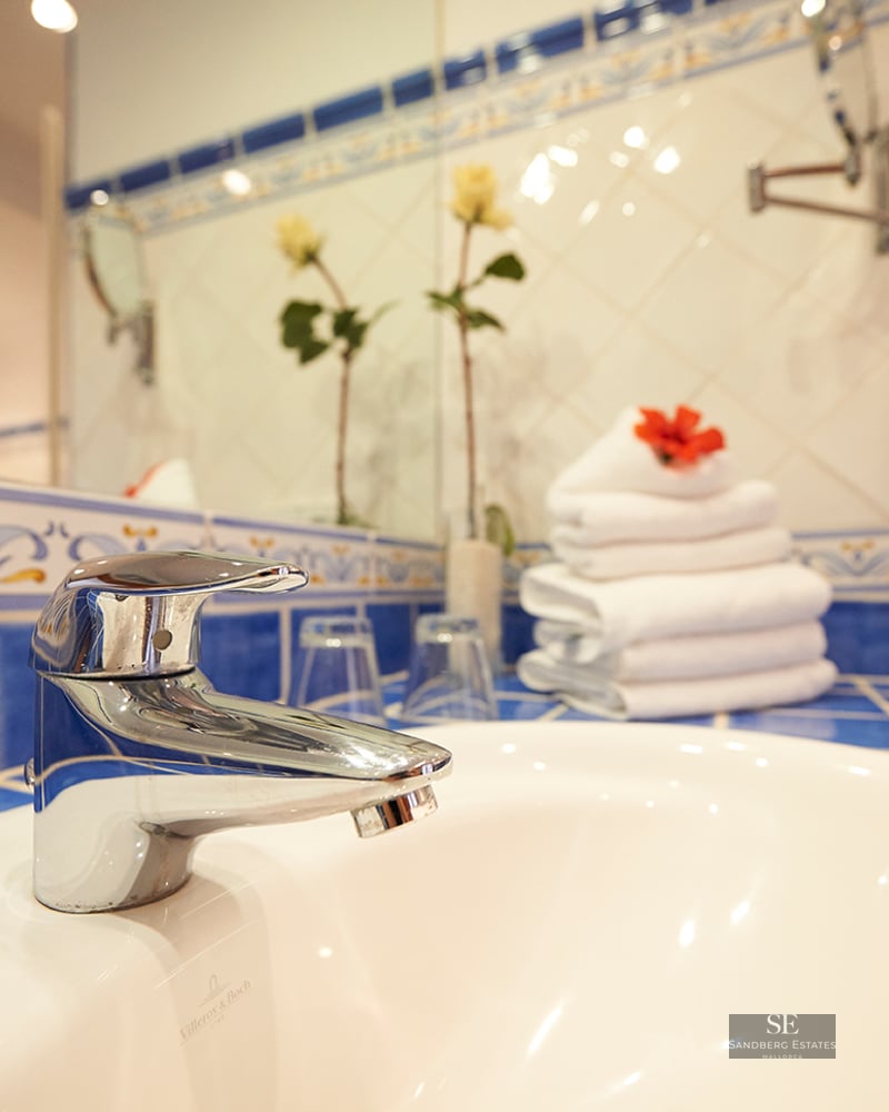 Close-up of a white porcelain sink and chrome faucet with blue patterned Mediterranean tiles and white towels.