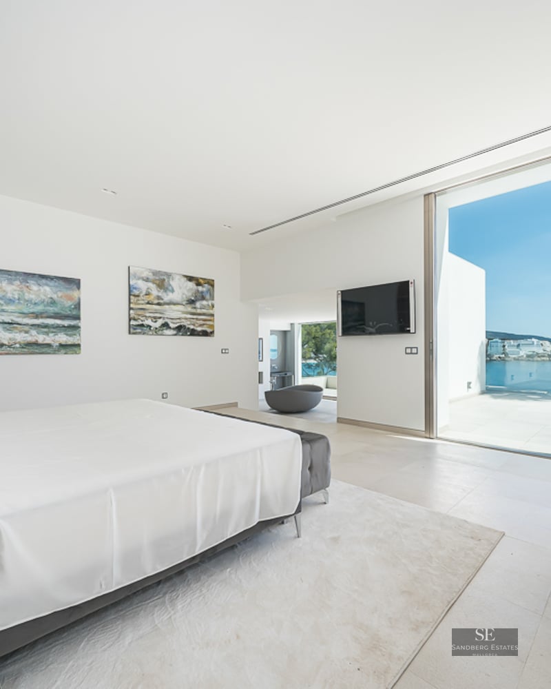 Bright master bedroom with white bedding, grey velvet chairs, and large glass doors overlooking the Mediterranean Sea.