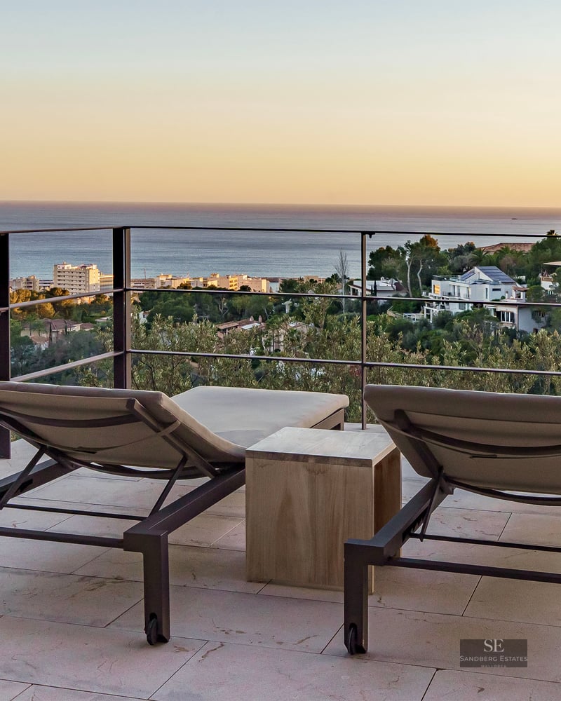Two modern sun loungers on a stone terrace facing a metal railing with panoramic sea and coastal views at sunset.