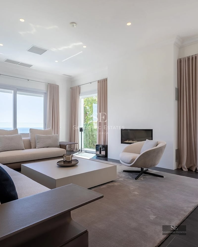 Spacious modern living room with beige sofas, a minimalist fireplace, and large windows overlooking the sea.