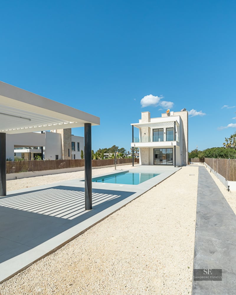 Exterior of a modern villa featuring a blue rectangular pool, white pergola, and gravel courtyard under a clear sky.