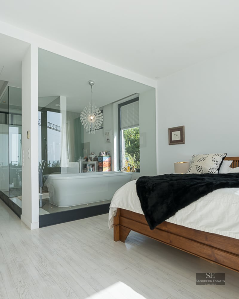 Modern master bedroom featuring a wooden bed and a glass-walled bathroom with a freestanding tub and designer chandelier.