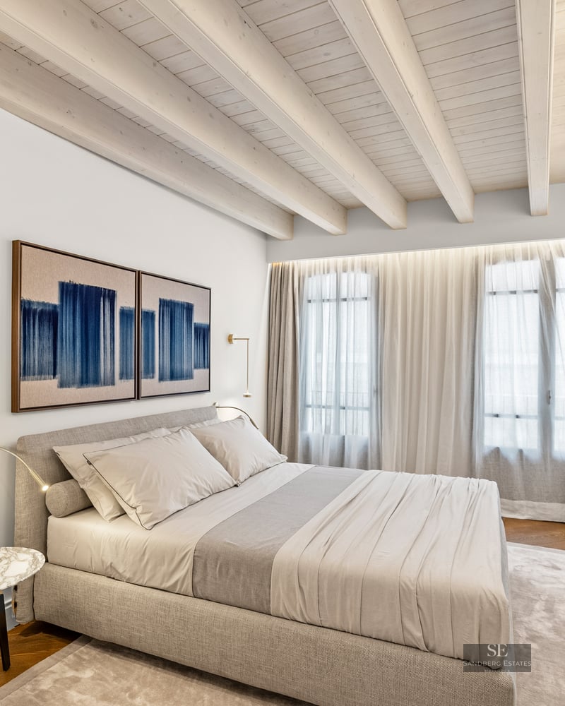 Modern bedroom with wooden ceiling beams, grey upholstered bed, marble side table, and floor-to-ceiling curtains.