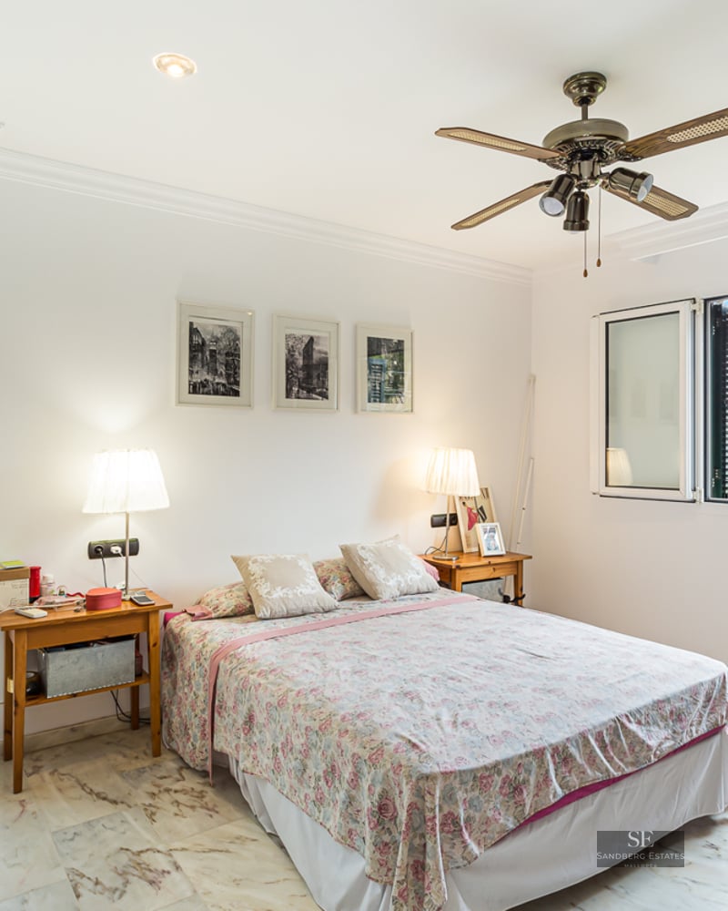 White bedroom with marble floor, double bed, ceiling fan, and window showing a scenic sea and forest view.