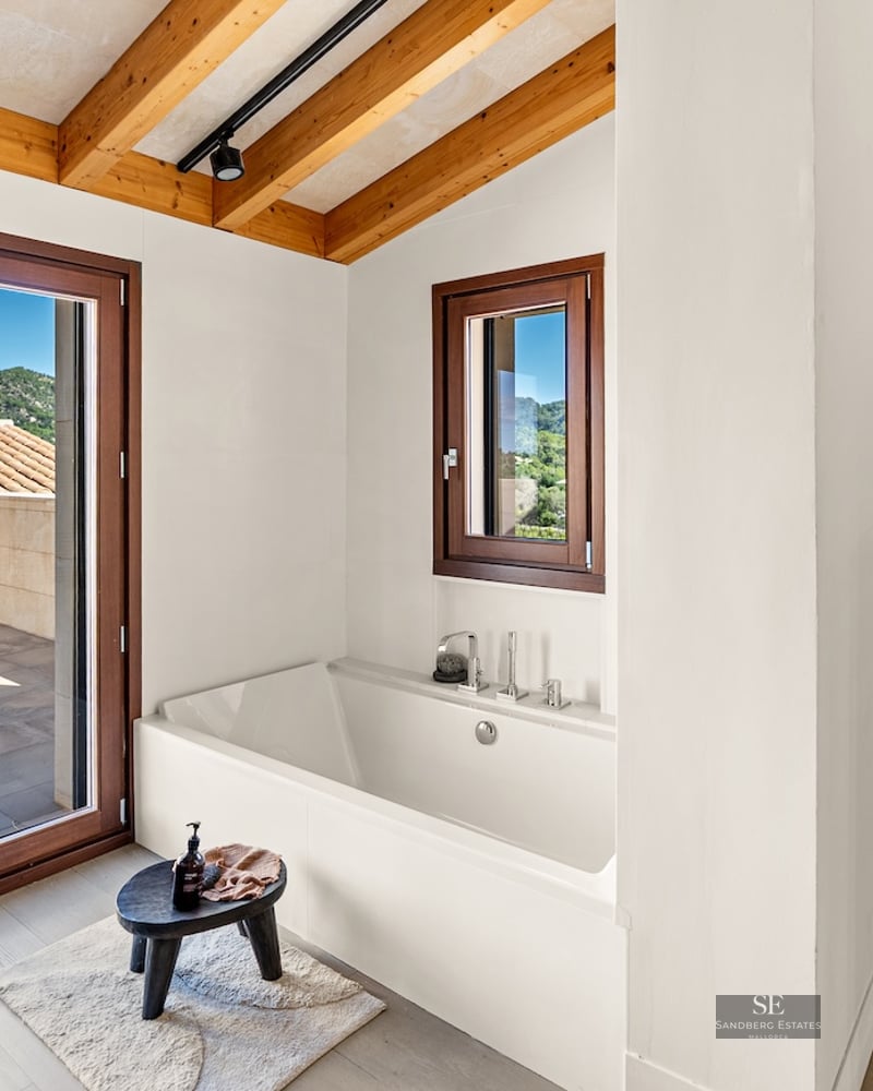 White built-in bathtub under a wooden beam ceiling with access to a terrace and mountain views.