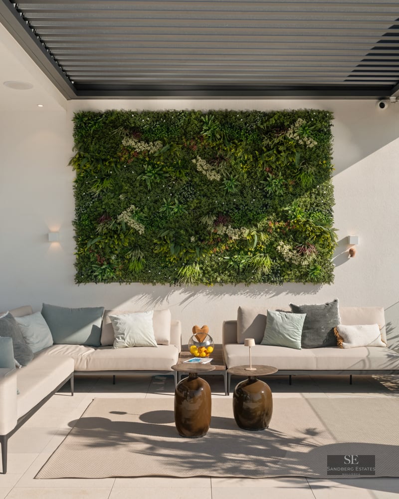 A contemporary outdoor terrace featuring a large beige corner sofa, a lush green vertical garden wall, and a louvered pergola.