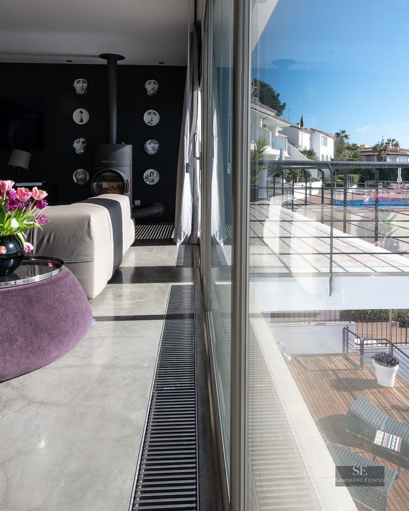 Modern living room with polished concrete floor, beige sofa, purple ottoman, and views of a sunny terrace through large glass doors.