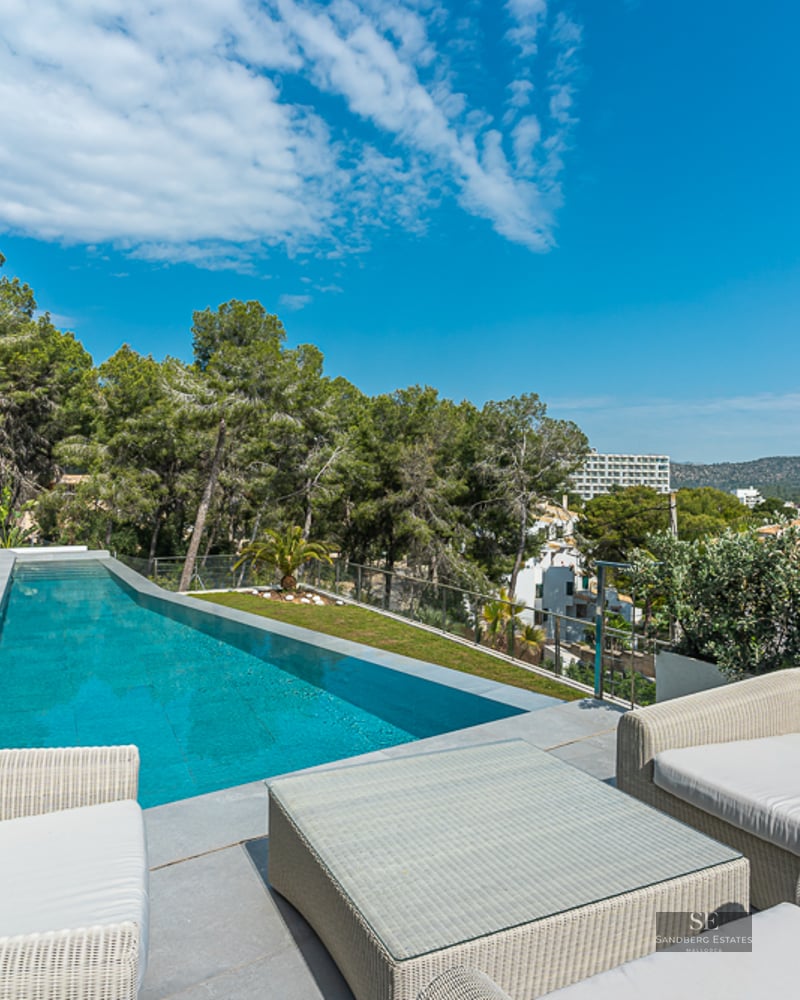 Modern terrace with a long blue swimming pool, white wicker lounge furniture, glass railings, and forest views.