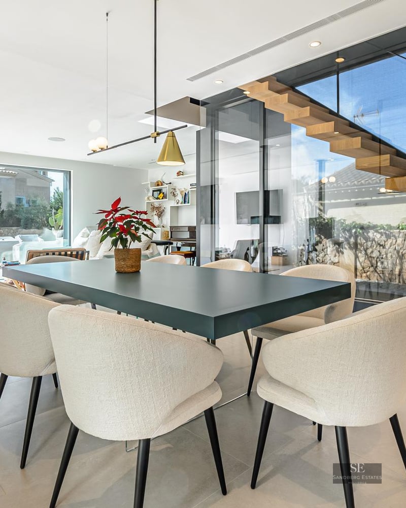 Contemporary dining room featuring a dark table, beige chairs, a floating wooden staircase, and large glass windows.