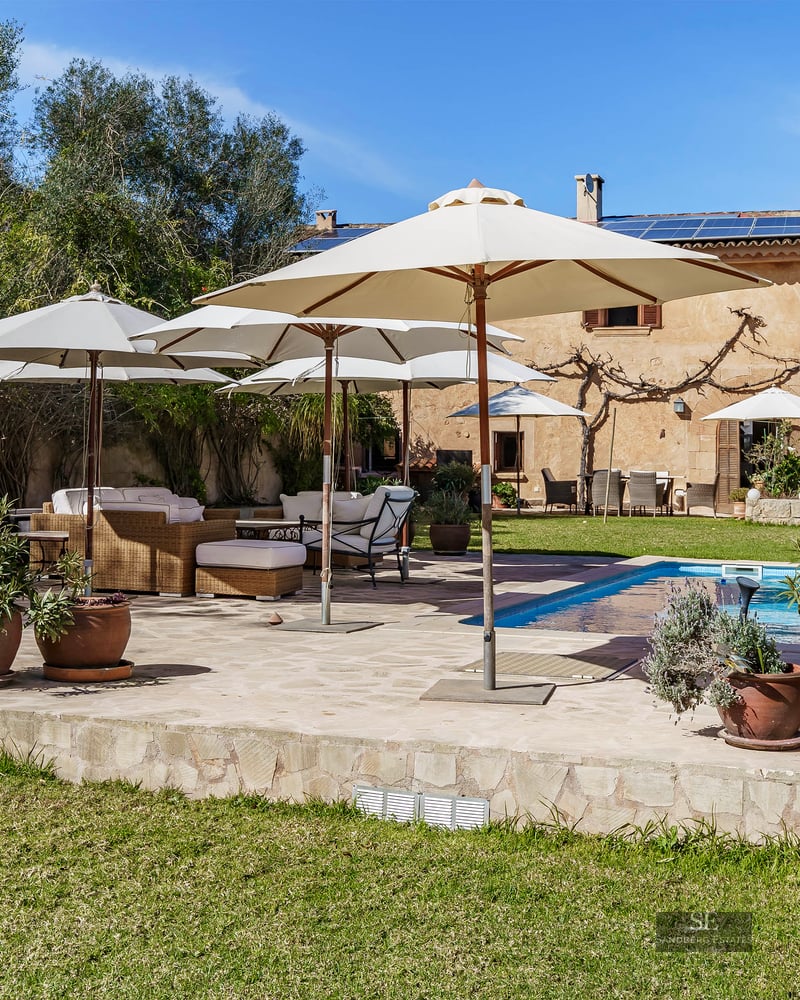 Large blue swimming pool surrounded by a stone deck with white umbrellas and wicker lounge furniture in a sunny garden.