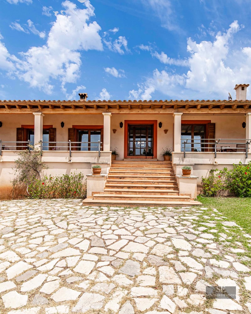 Front view of a one-story Mediterranean villa with a stone driveway, steps, and a covered terrace under a blue sky.