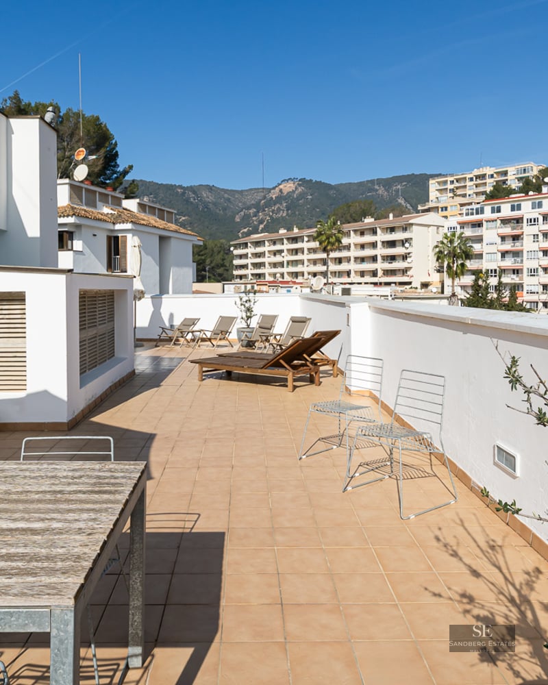 Large terrace with terracotta tiles, wooden sun loungers, and dining table under a clear blue sky.