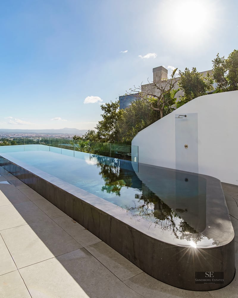 A sleek, long infinity pool on a minimalist white terrace overlooking a distant city under a bright blue sky.