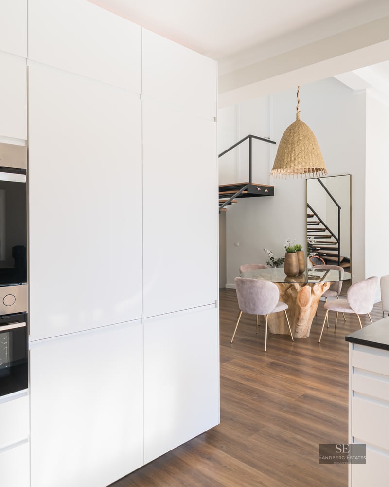 Cuisine blanche minimaliste avec fours encastrés donnant sur une salle à manger avec table en verre et chaises roses.