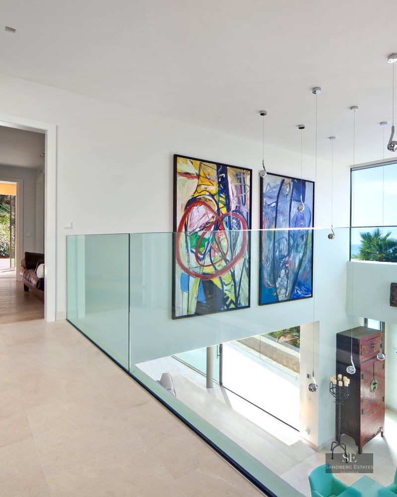 Modern hallway with glass railing, large paintings, and sea views through expansive windows.