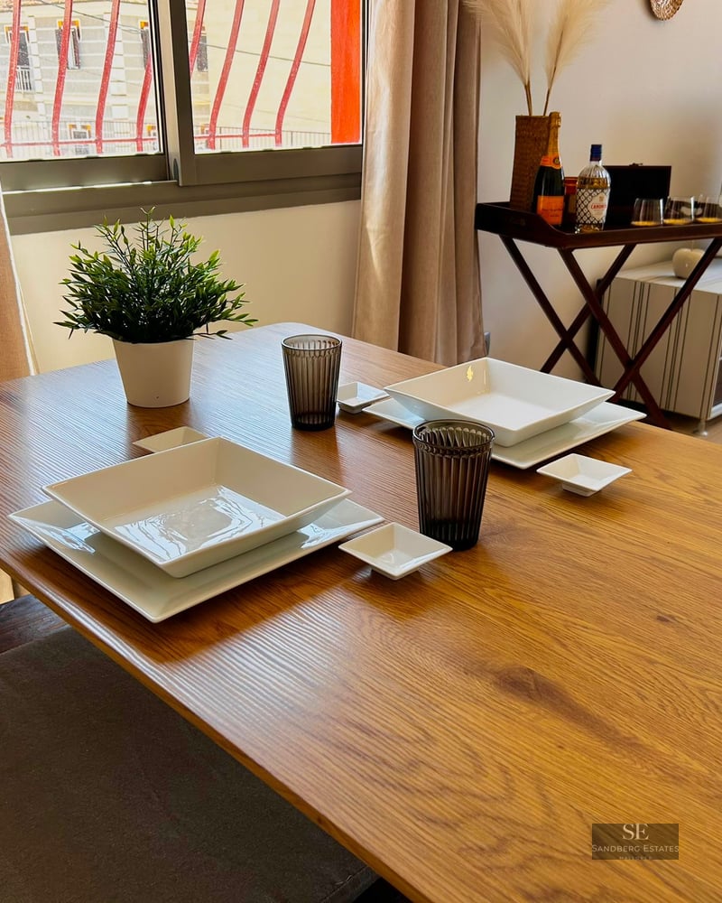 Wooden dining table set with white square plates and glasses in a bright living room.