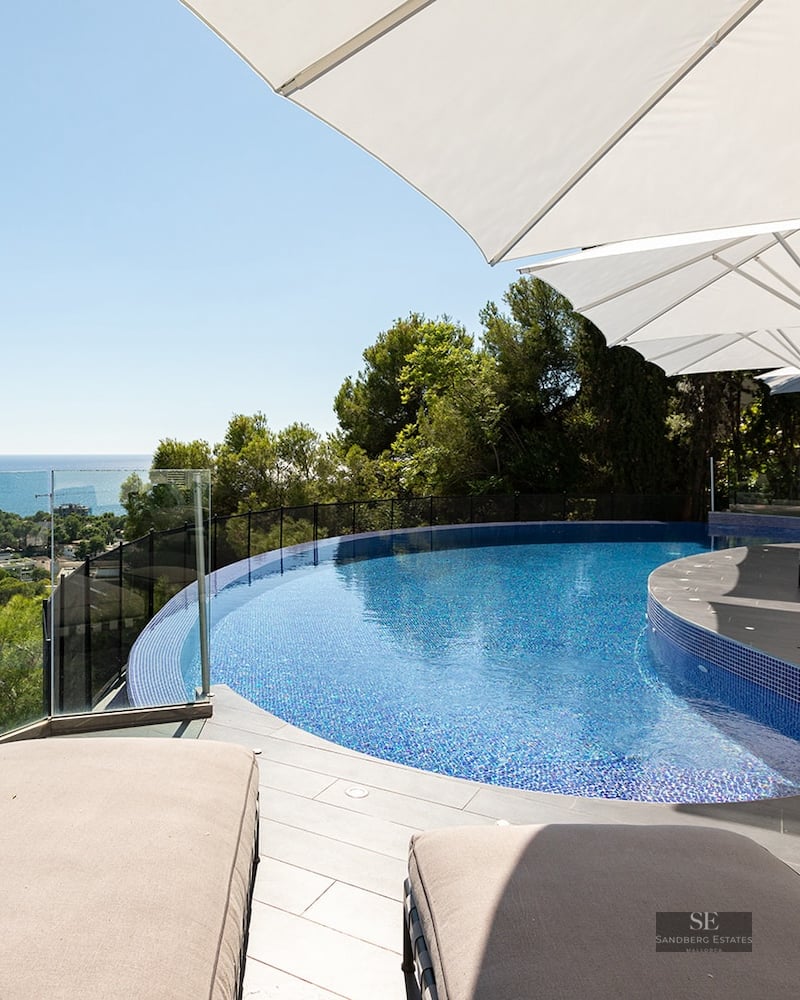 A curved blue swimming pool on a modern stone terrace with white umbrellas and sunbeds overlooking the sea.