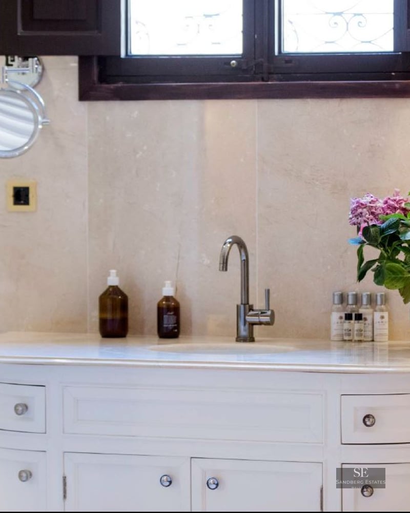 White vanity with chrome faucet, beige marble walls, magnifying mirror, and pink flowers in a pot.