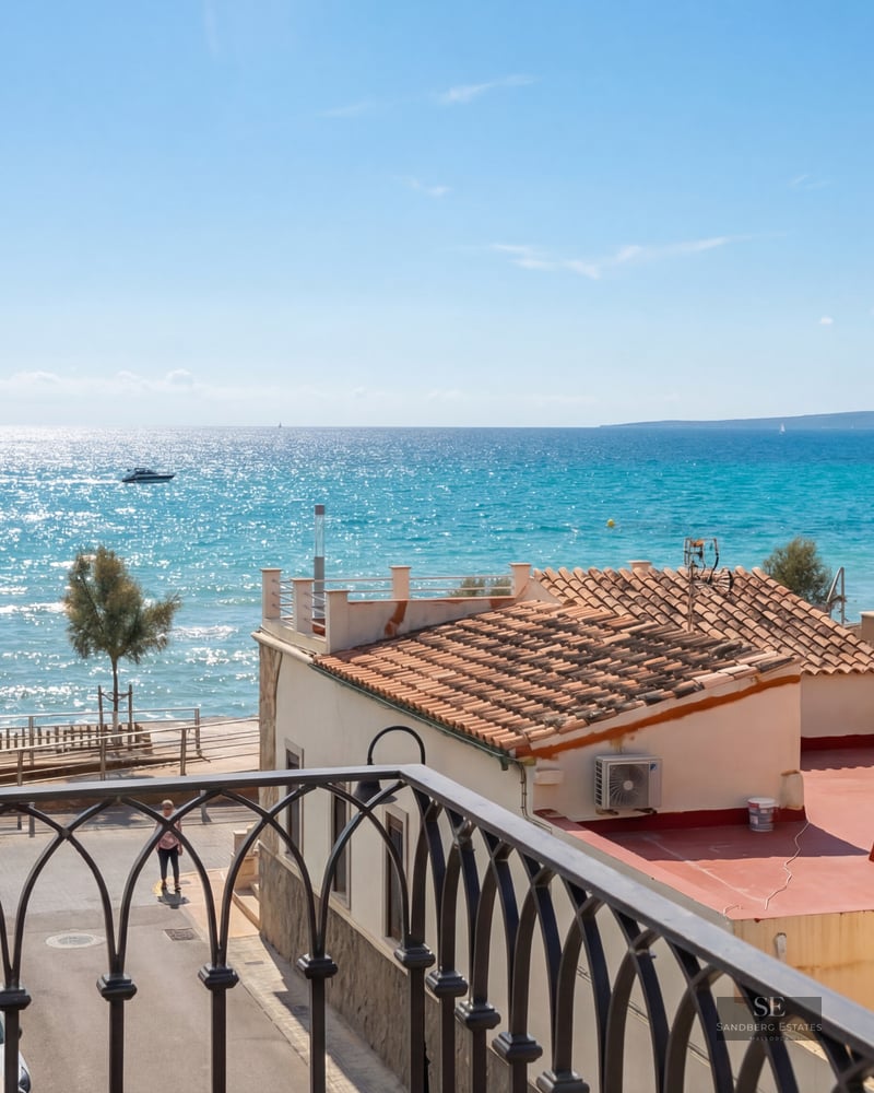 High-angle view from a wrought iron balcony looking out over terracotta rooftops toward a sparkling turquoise sea.