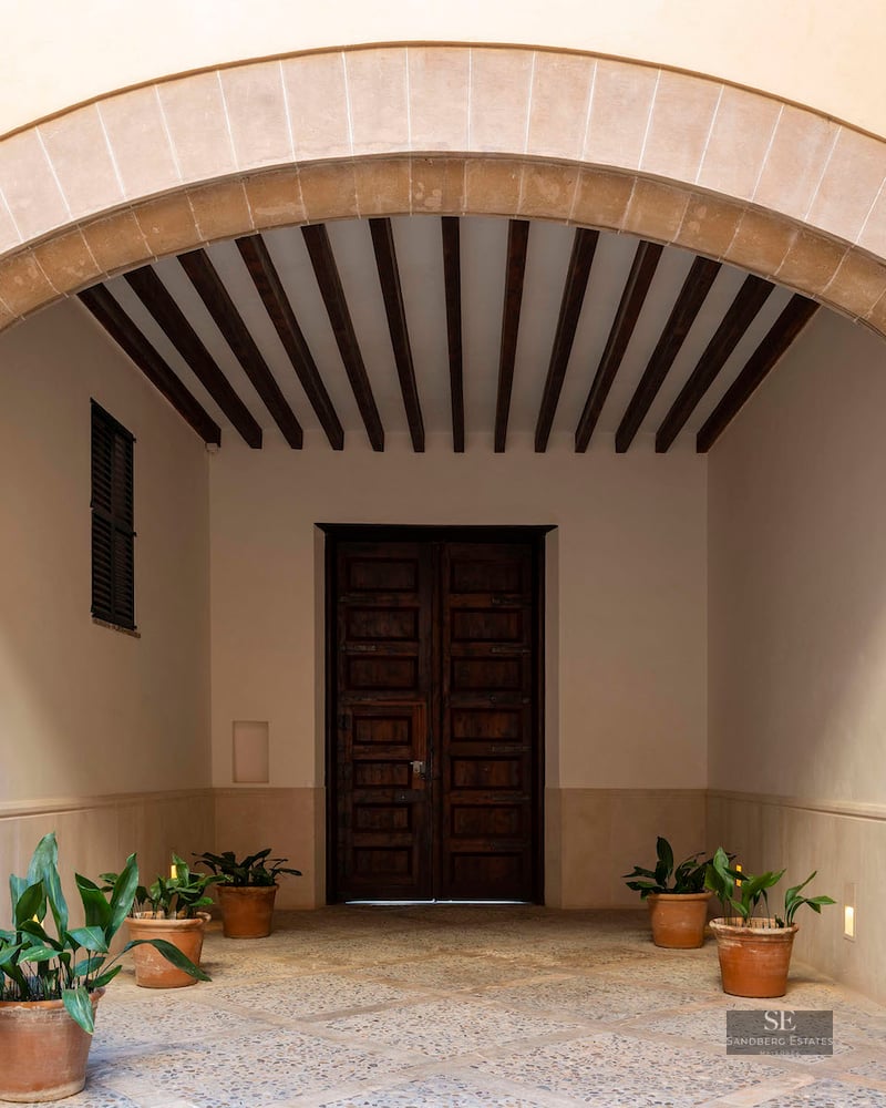 A grand stone archway leads to a traditional wooden door with exposed ceiling beams and potted plants.