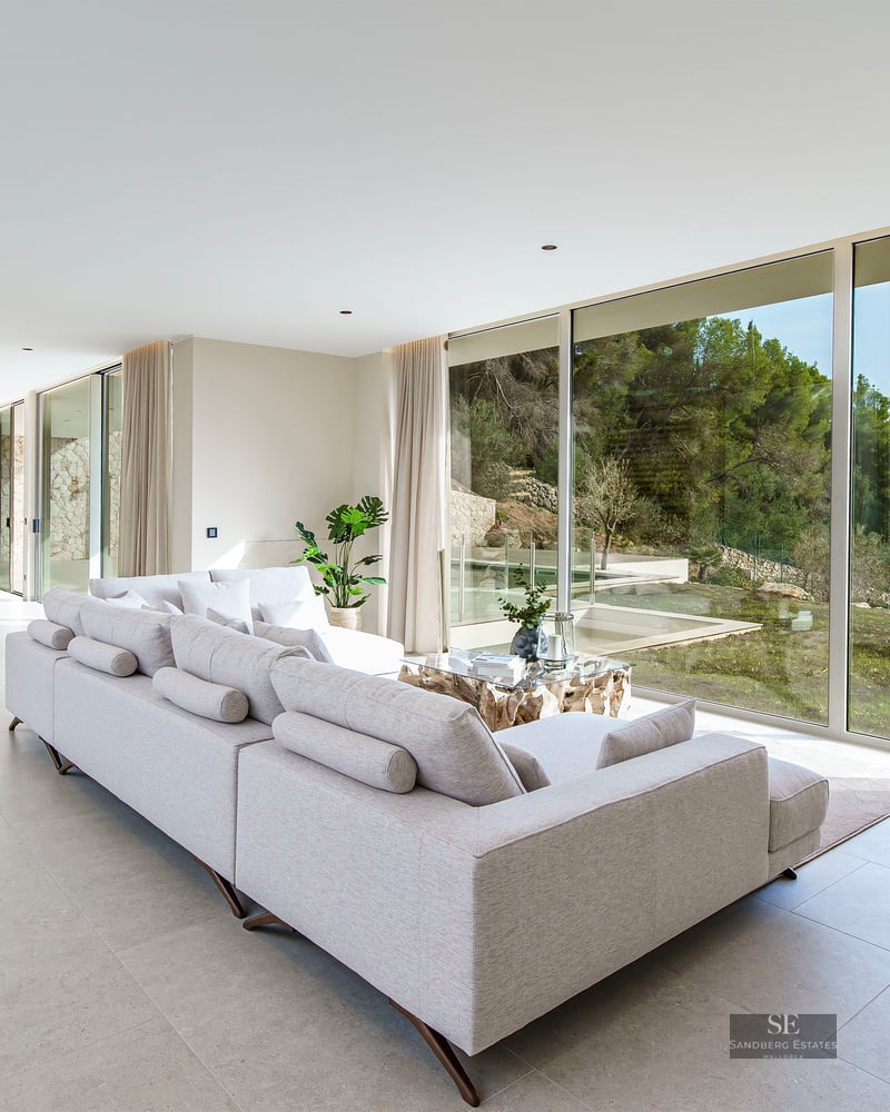 Spacious modern living room with a large grey sofa and floor-to-ceiling windows overlooking a lush green forest.