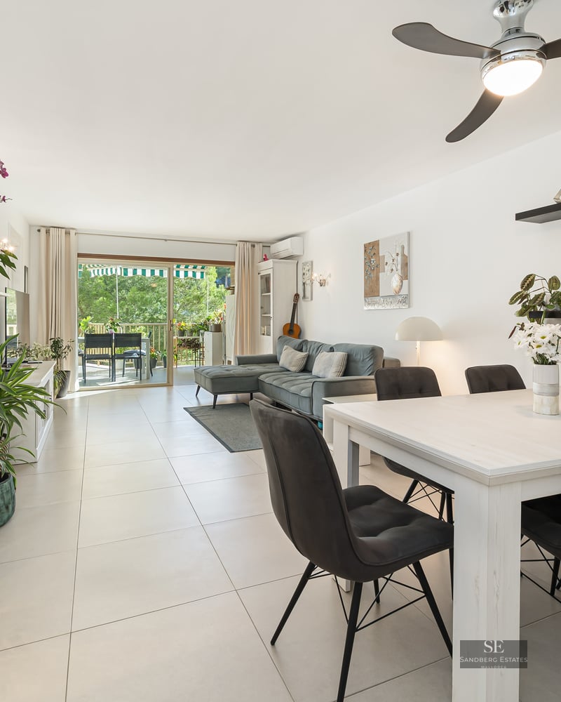 Espace de vie lumineux avec table à manger blanche, canapé gris et baies vitrées ouvrant sur un balcon verdoyant.