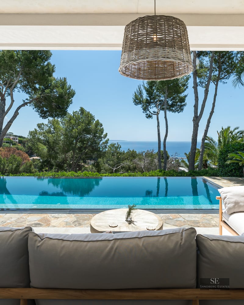 Infinity pool and outdoor lounge area under a shaded terrace overlooking the sea and lush green pine trees.