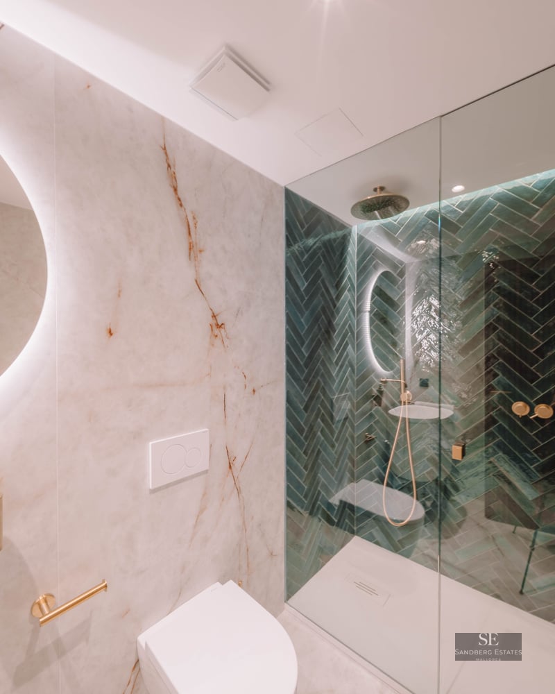 Modern bathroom with white marble walls, a round backlit mirror, and a walk-in shower with green herringbone tiles.