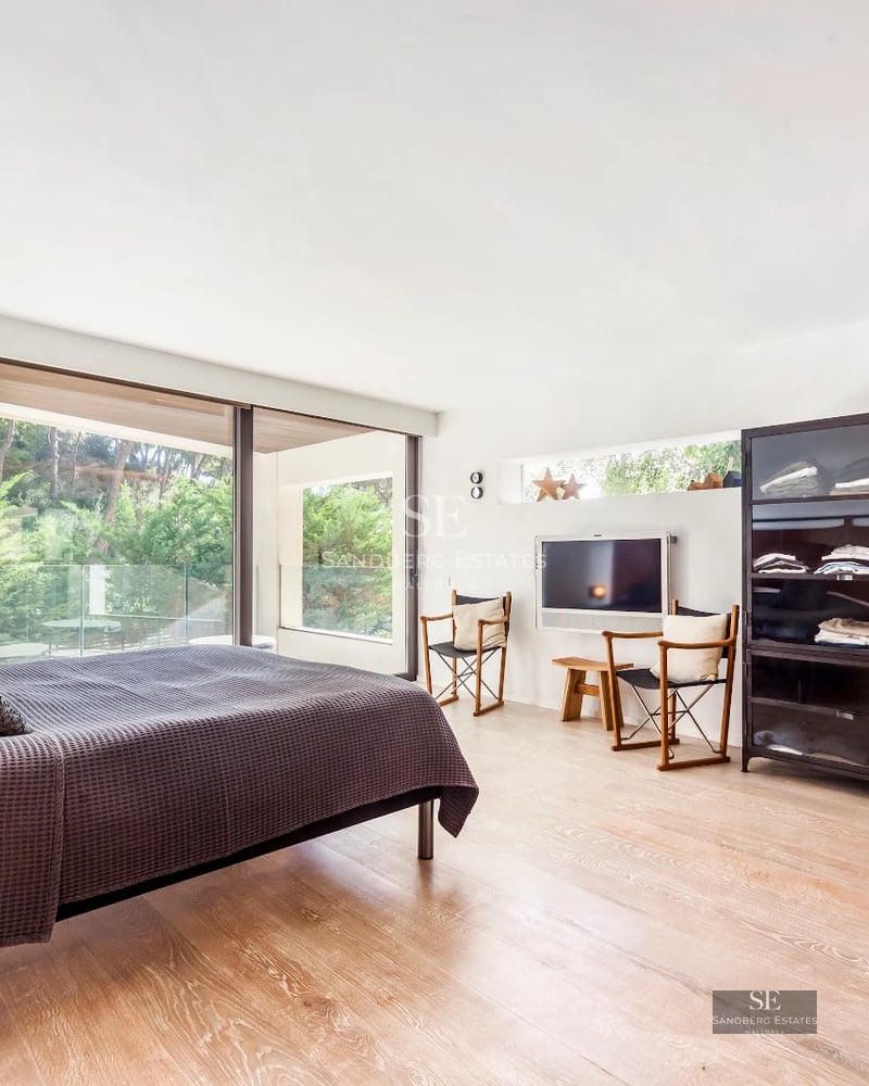 View of infinity pool with wood edge and sun loungers, surrounded by vegetation. Modern architecture with large windows.